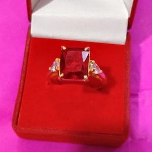 Op de afbeelding: Een gouden ring met een grote, rechthoekige, rode edelsteen als middelpunt. Twee kleinere, driehoekige, heldere edelstenen flankeren de hoofdsteen. De ring wordt gepresenteerd in een rode sieradendoos met een wit interieur.