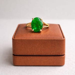 May include: A gold ring with a large, oval-cut emerald gemstone. The ring is displayed on a brown jewellery box. The emerald is a vivid green, and the gold band has a split shank design.