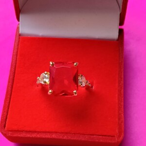 Op de afbeelding: Een gouden ring met een grote, rechthoekige rode edelsteen als middelpunt, geflankeerd door kleinere, heldere edelstenen. De ring wordt gepresenteerd in een rode fluwelen doos tegen een roze achtergrond, waarbij de sieraden worden uitgelicht.