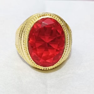 May include: A gold-toned ring featuring a large, oval-cut, faceted red gemstone set in the centre. The metal band displays a textured, decorative border surrounding the stone, with additional intricate detailing along the sides of the ring shank.