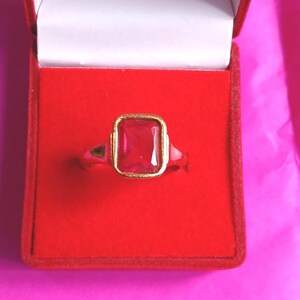 Op de afbeelding: Een goudkleurige ring met een grote, rechthoekige, rode edelsteen gezet in een gouden bezel. De ring wordt gepresenteerd in een rode fluwelen ringdoos met een witte binnenkant, tegen een roze achtergrond.