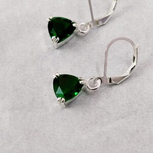 Puede incluir: Un par de pendientes de plata con cierre de palanca, que presentan piedras de color verde oscuro talladas en forma triangular y facetadas. Cada pendiente consta de un gancho de metal plateado pulido unido a una piedra verde engastada con garras.