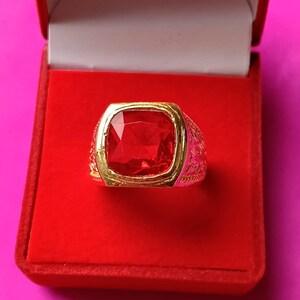 Peut inclure: Une bague dorée ornée d'une grande pierre précieuse rouge vif de forme carrée au centre. L'anneau de la bague présente des motifs floraux ou des volutes en relief complexes sur les côtés, donnant un aspect texturé au métal.