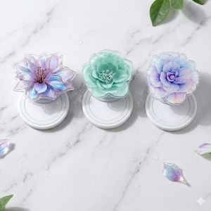 May include: Three floral phone grips in shades of purple, green, and blue. Each grip features a detailed flower design atop a white circular base. The flowers have translucent petals, creating a delicate, layered effect. The background is a marble-like surface.