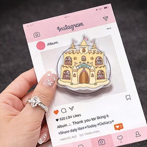 Op de afbeelding: Een doorzichtige telefoonhouder met een cartoon kasteelontwerp. Het kasteel is geel met bruine deuren en ramen, en het woord "BOOKSTORE" staat op de voorkant. De houder staat tegen een roze Instagram-thema achtergrond.