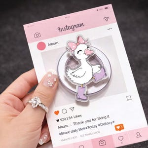 May include: A clear acrylic phone grip with a cartoon duck wearing purple wellington boots and a pink bow. The grip is set against a pink Instagram-themed background with the text "Instagram" and "Album."
