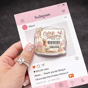 Op de afbeelding: Een heldere acryl telefoonhouder met de tekst "Cozy Readers Book Club" en illustraties van boeken, koffie en een vlinder. De houder staat op een roze Instagram-thema achtergrond.
