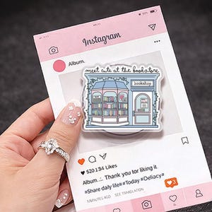 Op de afbeelding: Een sticker met een illustratie van een boekwinkel en de tekst "meet cute at the bookstore". Het staat tegen een roze Instagram-thema achtergrond. Een hand houdt de sticker vast, met een ring aan de vinger. De afbeelding heeft een social media thema.