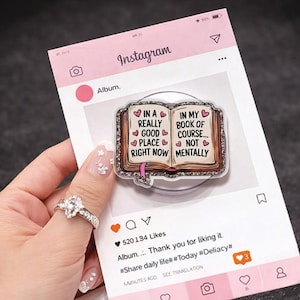 Op de afbeelding: Een doorzichtige telefoonhouder met een boekontwerp. Het boek heeft de tekst "IN A REALLY GOOD PLACE RIGHT NOW" aan de ene kant en "IN MY BOOK OF COURSE... NOT MENTALLY" aan de andere kant. De houder zit op een roze Instagram-kaart.