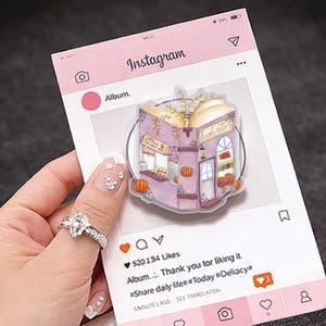 Op de afbeelding: Een doorzichtige popsocket met een paarse en witte illustratie van een koffiehuis en boekwinkel, versierd met pompoenen. De popsocket staat op een roze Instagram-achtergrond met de tekst "Instagram".