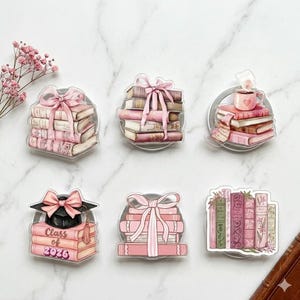 Puede incluir: Soportes transparentes para teléfono de acrílico con pilas de libros en tonos rosa y crema. Algunos tienen lazos rosas, un birrete o una taza con un corazón. El texto "Class of 2026" es visible en uno de ellos.
