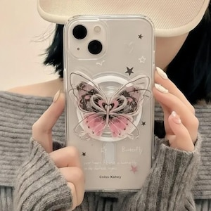 May include: Clear phone case with a butterfly design. The butterfly has pink and black wings. The case has a pop socket and is decorated with stars. The text "Butterfly" and "Cniss Kahey" are visible on the case.