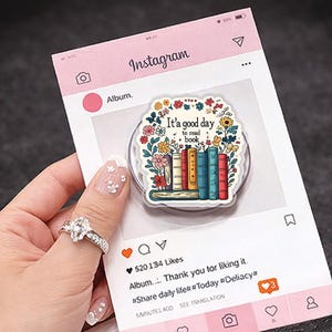 Op de afbeelding: Een doorzichtige telefoonhouder met een ontwerp van boeken en bloemen, met de tekst "It's a good day to read a book." De houder staat op een roze Instagram-thema achtergrond.