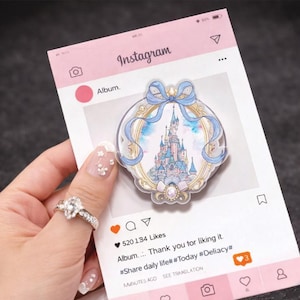 Puede incluir: Un bloc de notas rosa con temática de Instagram con un charm acrílico en forma de castillo. El charm presenta un marco azul y dorado con una pintura de acuarela de un castillo. El bloc de notas tiene el logotipo de Instagram y una leyenda que dice "Album... Thank you for liking it."