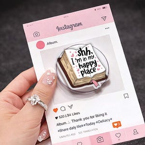 Op de afbeelding: Een roze Instagram-kaart met een sticker in de vorm van een boek met de tekst "shh, I'm in my happy place". De kaart heeft een roze en wit ontwerp met social media-iconen en tekst. De sticker is bruin, wit en roze.