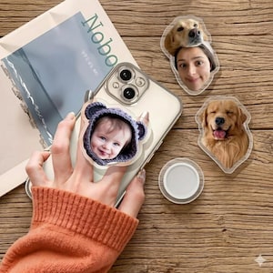May include: A clear phone case with a baby photo attached to a phone grip. Additional clear acrylic photo stickers of a woman and a golden retriever dog are also visible. The background includes a magazine and a wooden surface.