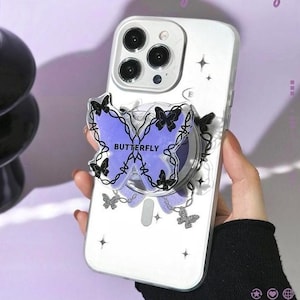 May include: A clear phone case featuring a purple butterfly design with the word "BUTTERFLY". The butterfly design has black accents and is attached to a white phone. The phone case has a pop-up grip with a butterfly design.