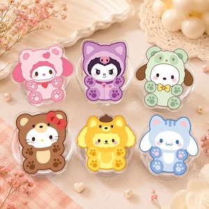 May include: Six colorful phone grips featuring cartoon characters in animal costumes. The grips include a pink bear, purple bat, green dog, brown bear, yellow dog, and blue cat. Each grip is round and has a clear base.