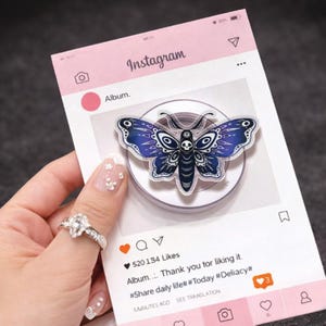 Puede incluir: Un soporte para teléfono con forma de mariposa azul y blanca. El soporte está sobre una base transparente y se muestra en una tarjeta rosa con temática de Instagram. La tarjeta tiene el logotipo de Instagram y el texto "Album... Gracias por gustarte."