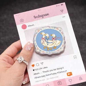 May include: A pop socket with a design of three white geese wearing bows, set on a light blue background with a floral border. The pop socket is displayed on a pink Instagram-themed card.