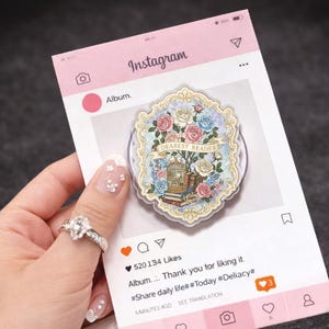May include: A decorative phone grip featuring a floral design with roses in shades of pink, blue, and white, and the text "DEAREST READER". The grip is held against a pink Instagram-themed background.