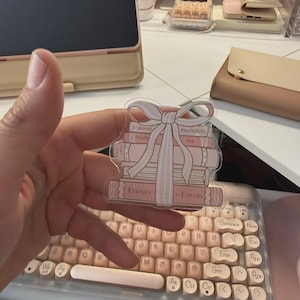 Op de afbeelding: Een doorzichtige sticker met een stapel roze boeken die met een lichtroze strik zijn vastgebonden. De boeken hebben titels als "Enemies to Lovers". De sticker wordt boven een roze en wit toetsenbord gehouden.