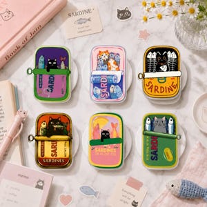 May include: Six rectangular sardine tin-shaped magnets or stickers featuring colorful illustrations of cats and fish. Each design displays a unique cat character interacting with sardines, with text including 'SARDINE' and 'IN OLIVE OIL' printed on the tins.