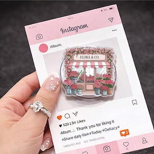 Op de afbeelding: Een roze telefoonhouder met Instagram-thema met een bloemenwinkelontwerp, met de tekst "FLORA & CO.". De houder is omgeven door roze bloemen en heeft een roze en wit gestreepte luifel. De Instagram-post toont likes en reacties.