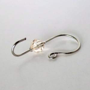 May include: A silver-toned metal ornament hook with a clear, faceted bead in the center. The hook has a curved design with a small loop on one end and a slightly open hook on the other. The bead adds a touch of sparkle.
