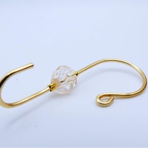 May include: A gold-colored metal hook with a clear, faceted bead in the center. The hook has a curved design with a loop on one end and an open end on the other. The bead adds a decorative element to the hook.