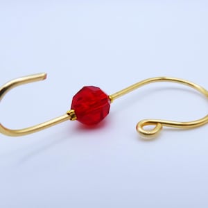 May include: A gold-colored earring hook with a faceted red bead in the center. The hook has a curved design with a loop on one end and an open hook on the other. The earring is likely for pierced ears.