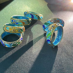 May include: A collection of ornate rings with a blue enamel interior and gold-coloured metal bands. The rings feature colourful floral designs in shades of blue, green, orange, and yellow. The rings are displayed on a dark green surface.