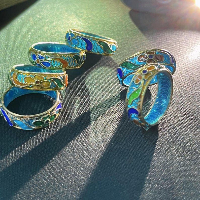 May include: A collection of ornate rings with a blue enamel interior and gold-coloured metal bands. The rings feature colourful floral designs in shades of blue, green, orange, and yellow. The rings are displayed on a dark green surface.