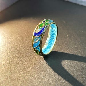 May include: A gold ring with detailed enamel work. The ring showcases a pattern of blue and green enamel, with a light blue interior. The design incorporates floral and abstract shapes, resulting in a unique and artistic piece of jewellery.