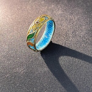May include: A detailed, vintage ring with a floral design. The ring features a gold-coloured band with colourful enamel accents in yellow, green, and orange. The interior of the ring is a bright blue. The ring is approximately 0.6 cm wide.