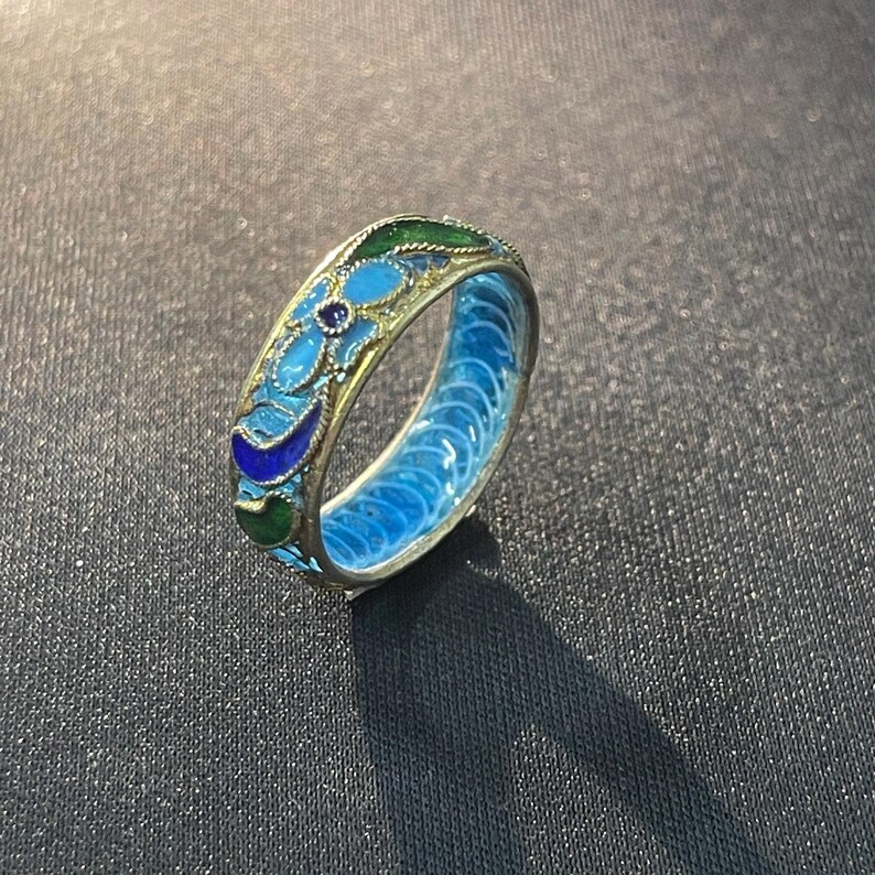 May include: A detailed, colourful ring with a floral design. The ring features a light blue interior and is adorned with blue, green, and gold accents. A small, dark blue stone is set in the centre of the floral pattern. The ring is set against a dark background.