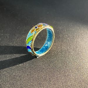 May include: A gold ring with detailed enamel work. The ring has a colourful design with blue, green, orange, and yellow enamel. The inside of the ring is bright blue. The ring is set against a dark background.