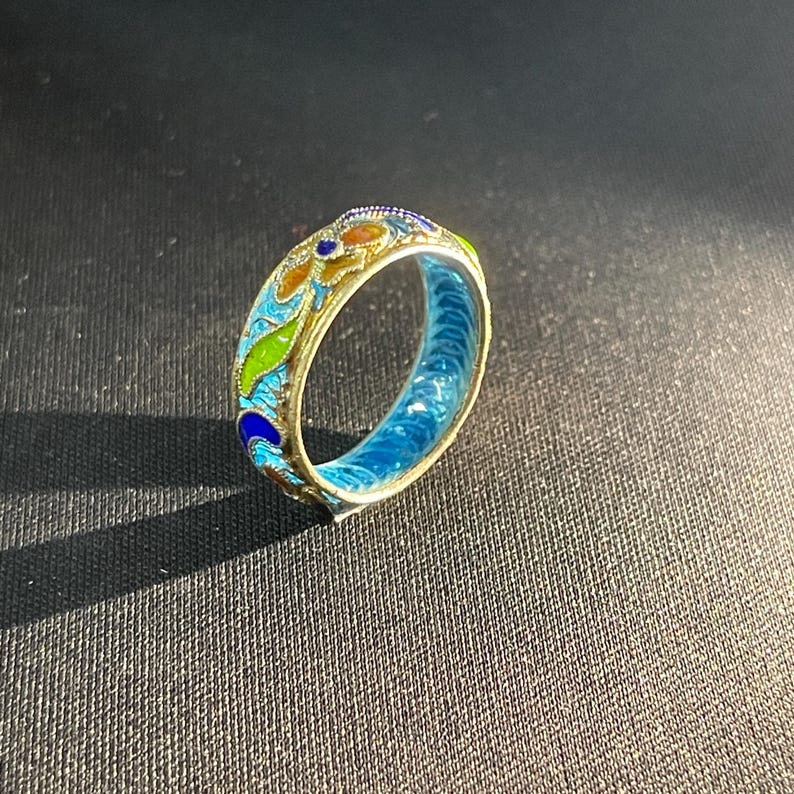 May include: A gold ring with detailed enamel work. The ring has a colourful design with blue, green, orange, and yellow enamel. The inside of the ring is bright blue. The ring is set against a dark background.