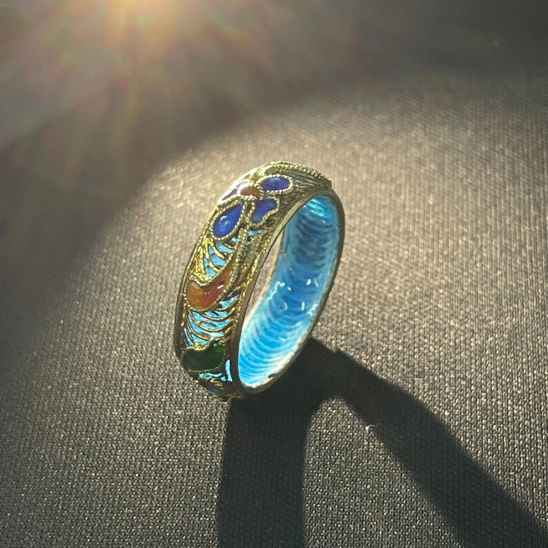 May include: A gold-coloured ring featuring a floral pattern in blue, orange, and green enamel. The inside of the ring is bright blue. The ring is set against a dark background, with sunlight shining on the ring.