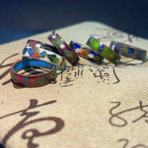 May include: A collection of colorful enamel rings with floral designs. The rings feature a variety of colors, including red, blue, green, and white, with intricate patterns. The rings are displayed on a textured, tan surface.