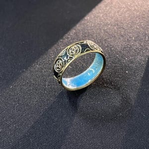 May include: A gold ring with a black enamel band displaying a repeating floral pattern. The inside of the ring is light blue. The ring is set against a dark background.