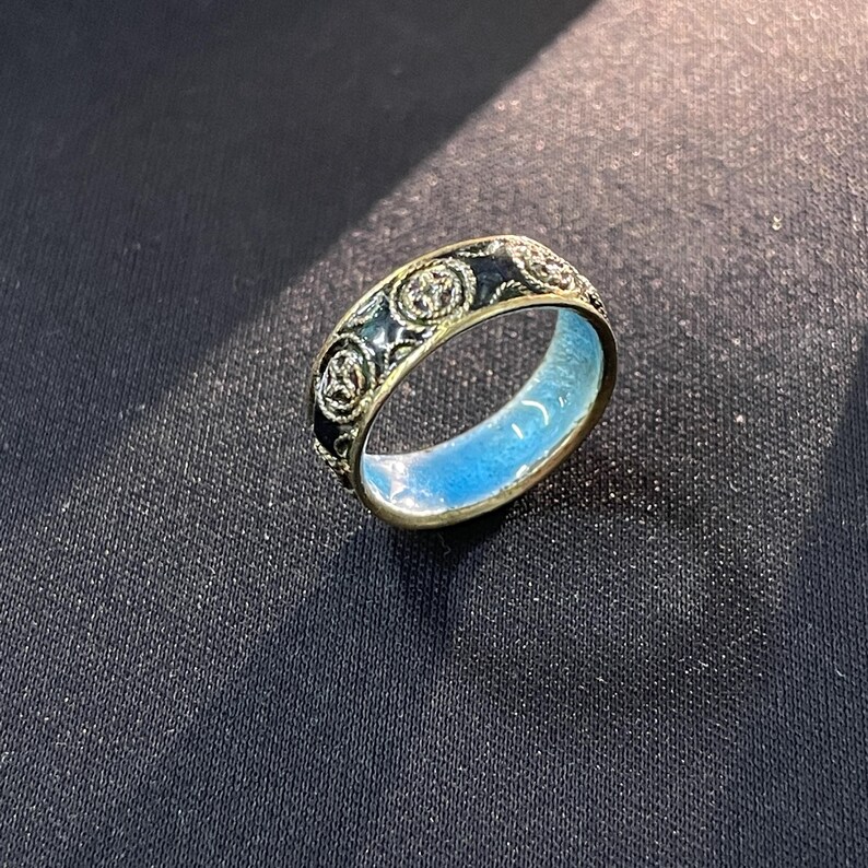 May include: A gold ring with a black enamel band displaying a repeating floral pattern. The inside of the ring is light blue. The ring is set against a dark background.