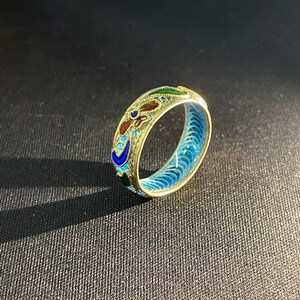May include: A gold ring with detailed enamel work. The ring has a floral design with red, blue, green, and gold colours. The inside of the ring is bright blue. The ring is displayed against a dark backdrop.