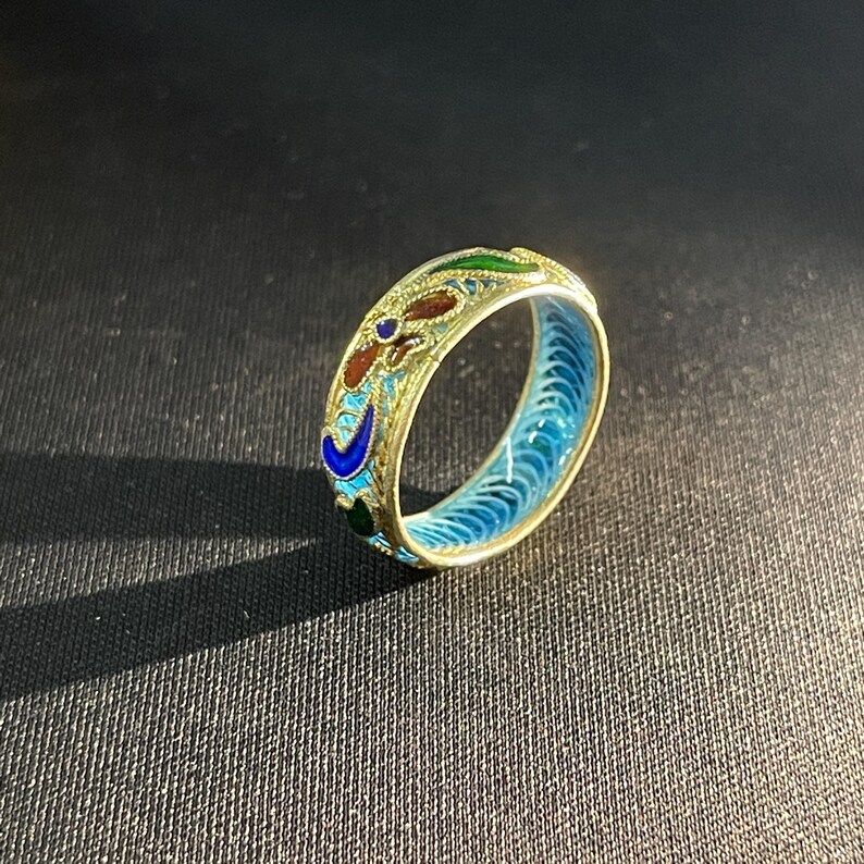 May include: A gold ring with detailed enamel work. The ring has a floral design with red, blue, green, and gold colours. The inside of the ring is bright blue. The ring is displayed against a dark backdrop.