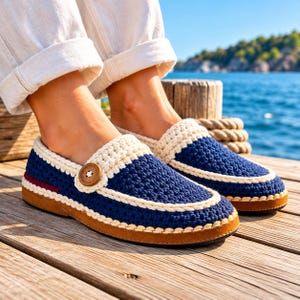 May include: A pair of navy blue crochet loafers featuring a cream-coloured trim, a single brown wooden button on the side, and a flat tan sole. The shoes are worn with white cuffed trousers, showcasing a textured knit construction.