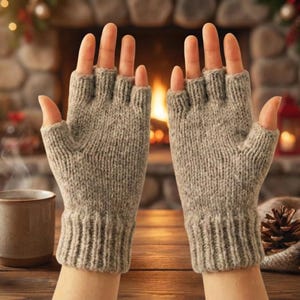 May include: Light grey fingerless gloves made from knitted material. The gloves are shown against a warm, cosy backdrop with a fireplace, a mug of coffee, and pine cones, suggesting a winter theme.