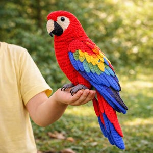 May include: A handcrafted, crochet parrot in vibrant red, blue, yellow, and green. The bird has a black beak, gray feet, and is perched on a hand. The parrot's wings and tail feature a spectrum of colors, creating a striking visual appeal.