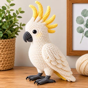 May include: A crocheted cockatoo figurine featuring a white body, a bright yellow crest, a dark gray beak, and dark gray feet. The bird has black bead eyes and is crafted with visible yarn stitches, showcasing a detailed texture throughout.