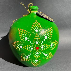 Puede incluir: Adorno redondo verde pintado a mano, con una cuerda dorada para colgar. Presenta un diseño de puntos blancos, amarillos y naranjas, con un punto rojo en el centro. Ideal para decoraciones festivas o de temporada.