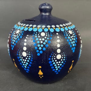 Puede incluir: Una hucha de cerámica azul marino con una ranura en la parte superior. Presenta un patrón de puntos blancos, azul claro y azul oscuro, con detalles dorados. La hucha tiene un pequeño pomo en la parte superior.